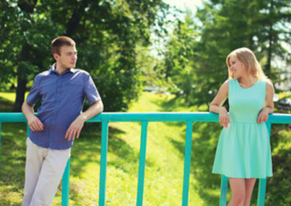Парень и девушка стоят на мосту поодаль и смотрят в глаза друг другу. Девушка улыбается