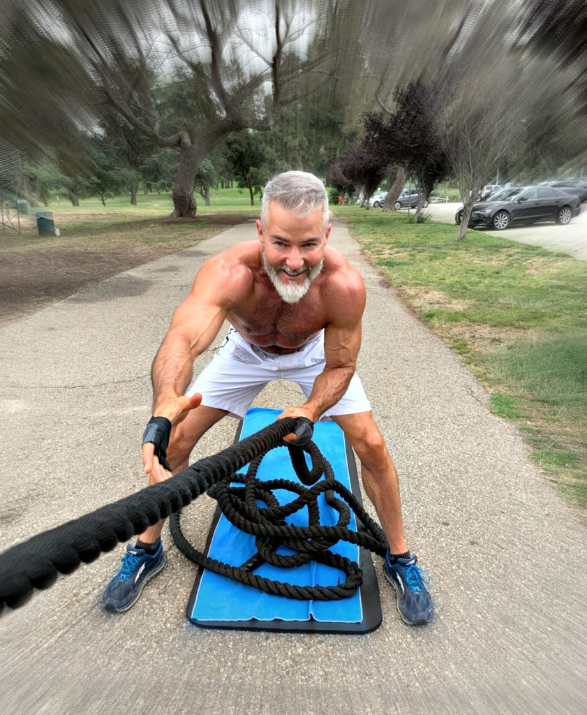 Muscled, older athlete demonstrates standing facing pulls with a battle rope, outdoors.