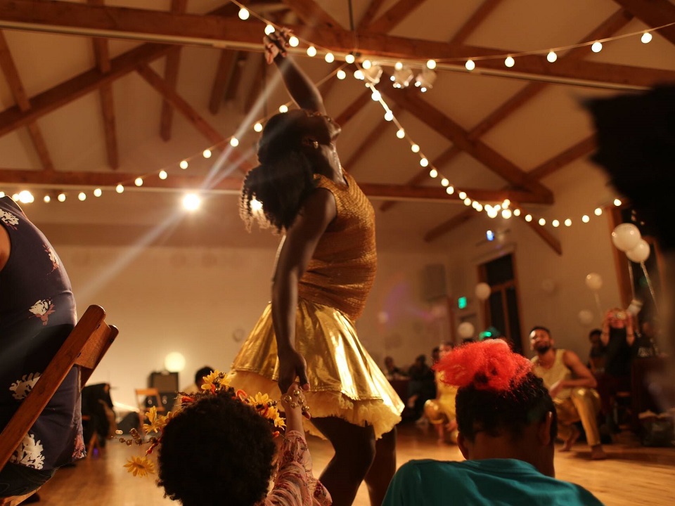 J'nae dressed in a mustard top and gold skirt, looking up and reaching high with one hand as dangling ceiling lights twinkle and seated audience watches